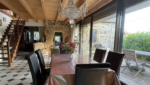 A dining area with a wooden table, black chairs, and a chandelier. There is a stone wall and a view of greenery outside. - BRITTANY FINISTERE CROZON PENTY SPACIOUS AND LUMINOUS (Crozon)