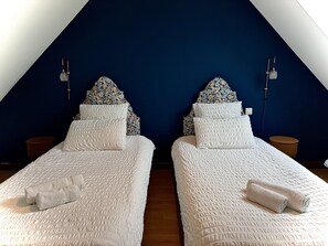 Dos camas individuales con ropa de cama blanca y almohadas estampadas, frente a una pared azul oscuro con techo inclinado.