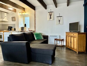 Una sala moderna con un sofá, un televisor sobre un mueble de madera, y una zona de cocina con lavavajillas y cafetera.