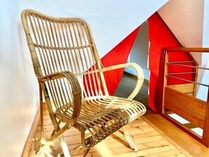 Ein Korbstuhl mit hoher Rückenlehne und Armlehnen, platziert in einer Ecke eines Raumes mit Holzboden und einer rot-beigen geometrischen Wandgestaltung.