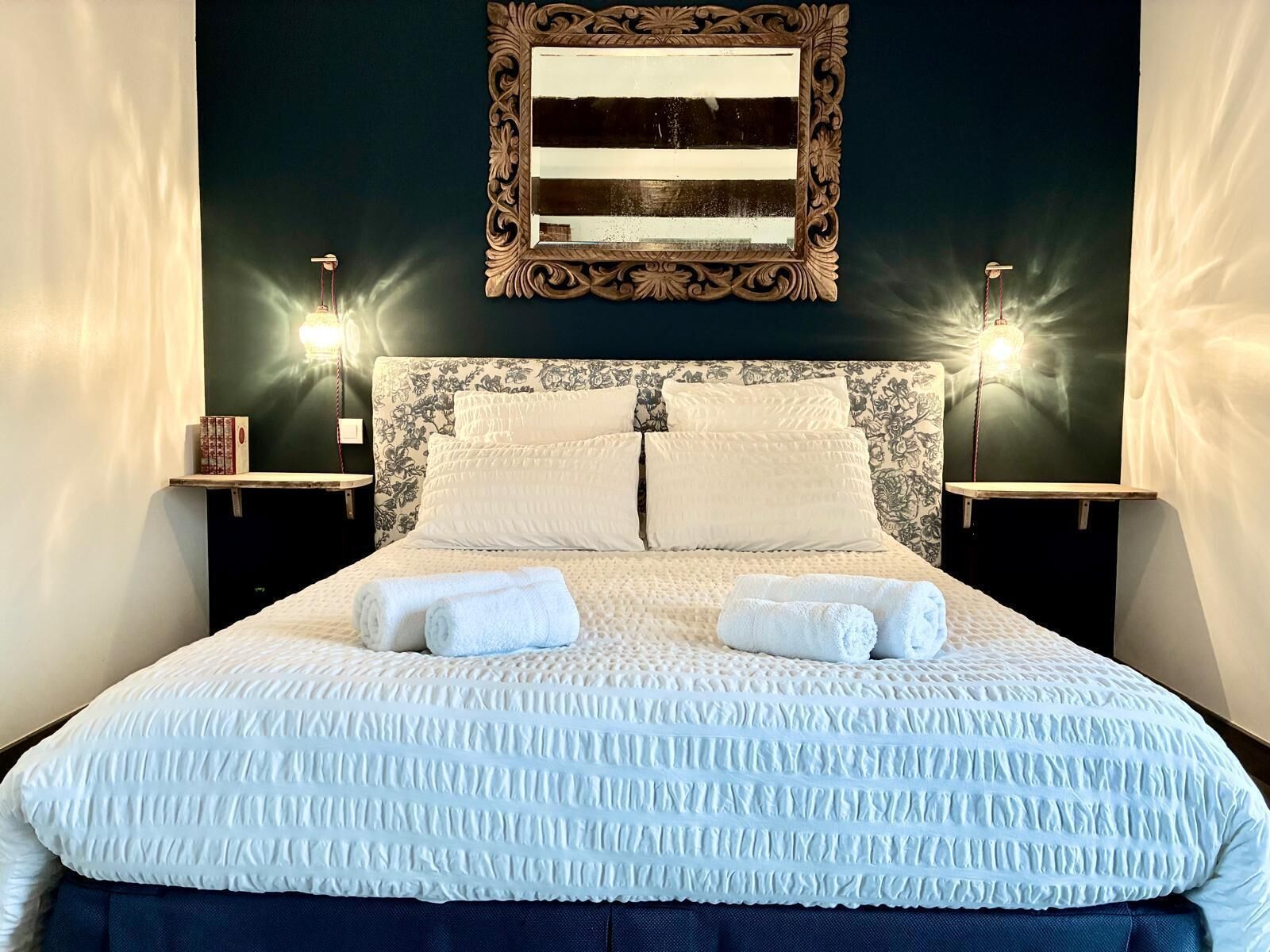 Cama bem arrumada com colcha estampada e almofadas, com duas mesas de cabeceira com abajures de cada lado, diante de uma parede escura com um espelho decorativo acima.