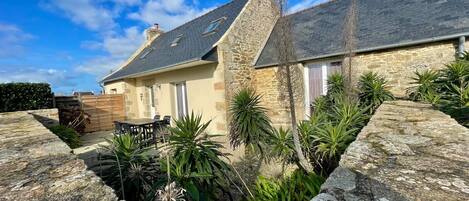 A stone house with a sloped roof, a stone wall, and a garden with various plants.