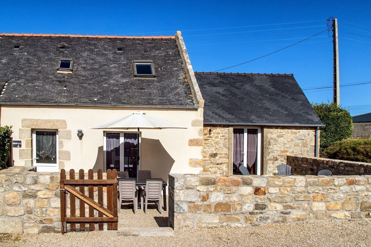 Un bâtiment en pierre avec un toit en pente, une porte en bois et une terrasse aménagée avec du mobilier d’extérieur.