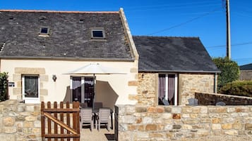 Un bâtiment en pierre avec un toit en pente, une porte en bois et une terrasse aménagée avec du mobilier d’extérieur.