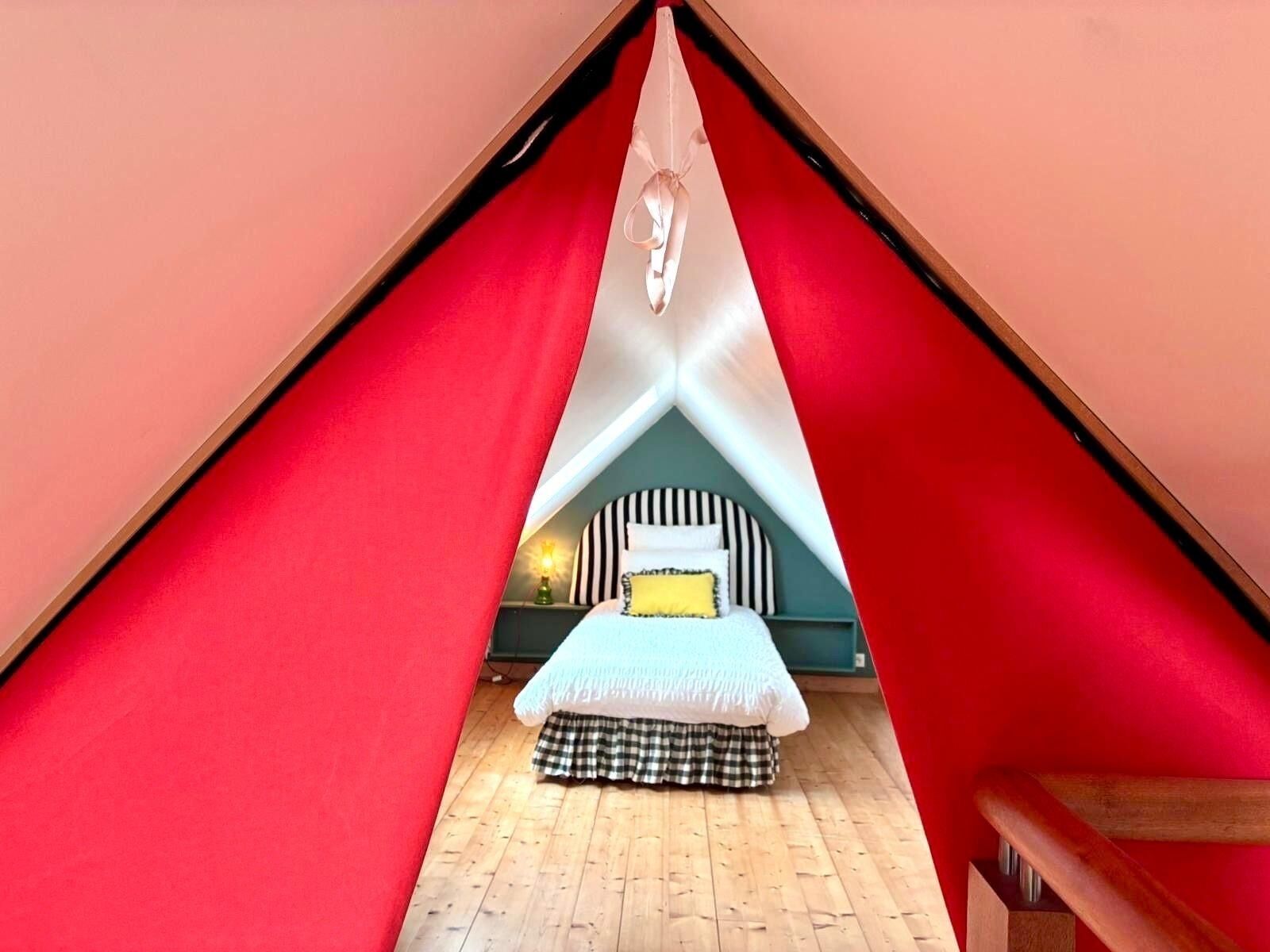 Une pièce triangulaire avec un lit au centre, une petite table avec une lampe et une chaise en bois.