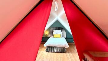 Une pièce triangulaire avec un lit au centre, une petite table avec une lampe et une chaise en bois.