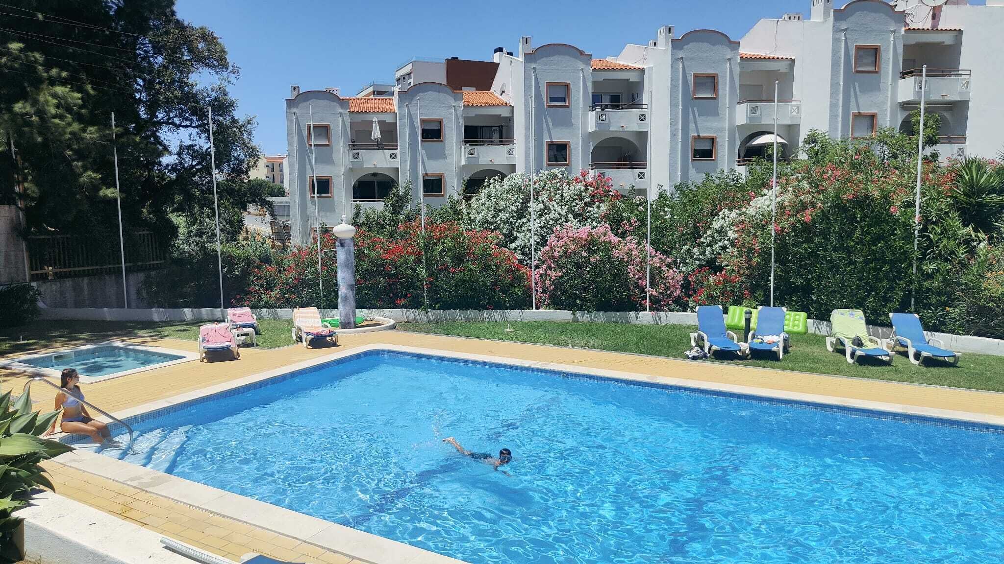 A swimming pool with a person swimming, surrounded by lounge chairs and a building with balconies.