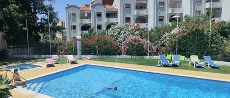 A swimming pool with a person swimming, surrounded by lounge chairs and a building with balconies.