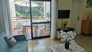A living room with a sofa, a coffee table with a vase of flowers, a TV on the wall, and a sliding glass door leading to an outdoor patio with a table and chairs.