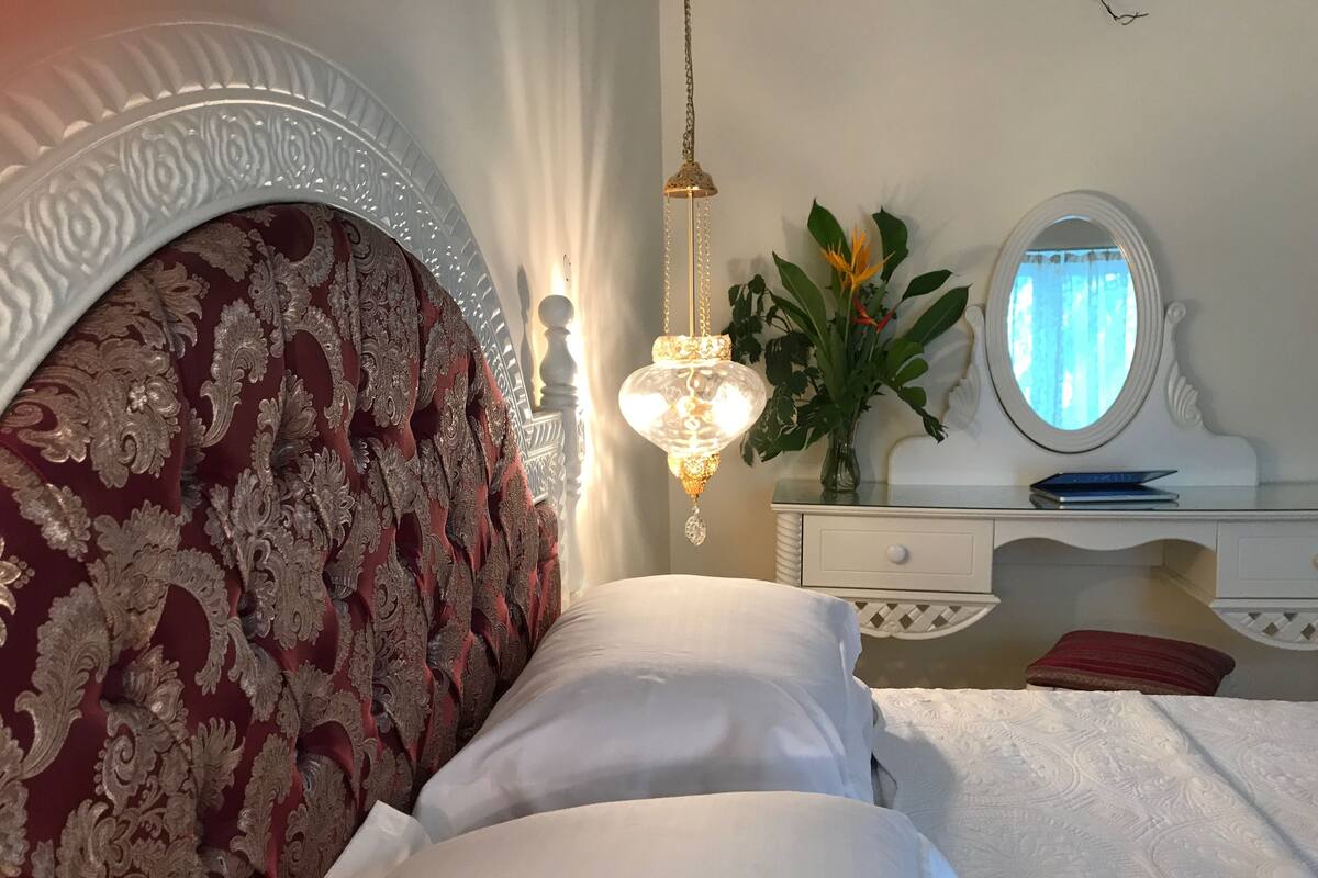 Une chambre à coucher avec une grande tête de lit décorative, une table de chevet avec un miroir, une lampe et une plante.