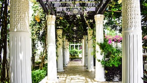 Un chemin de jardin couvert, bordé de colonnes blanches et entouré de verdure et de fleurs.