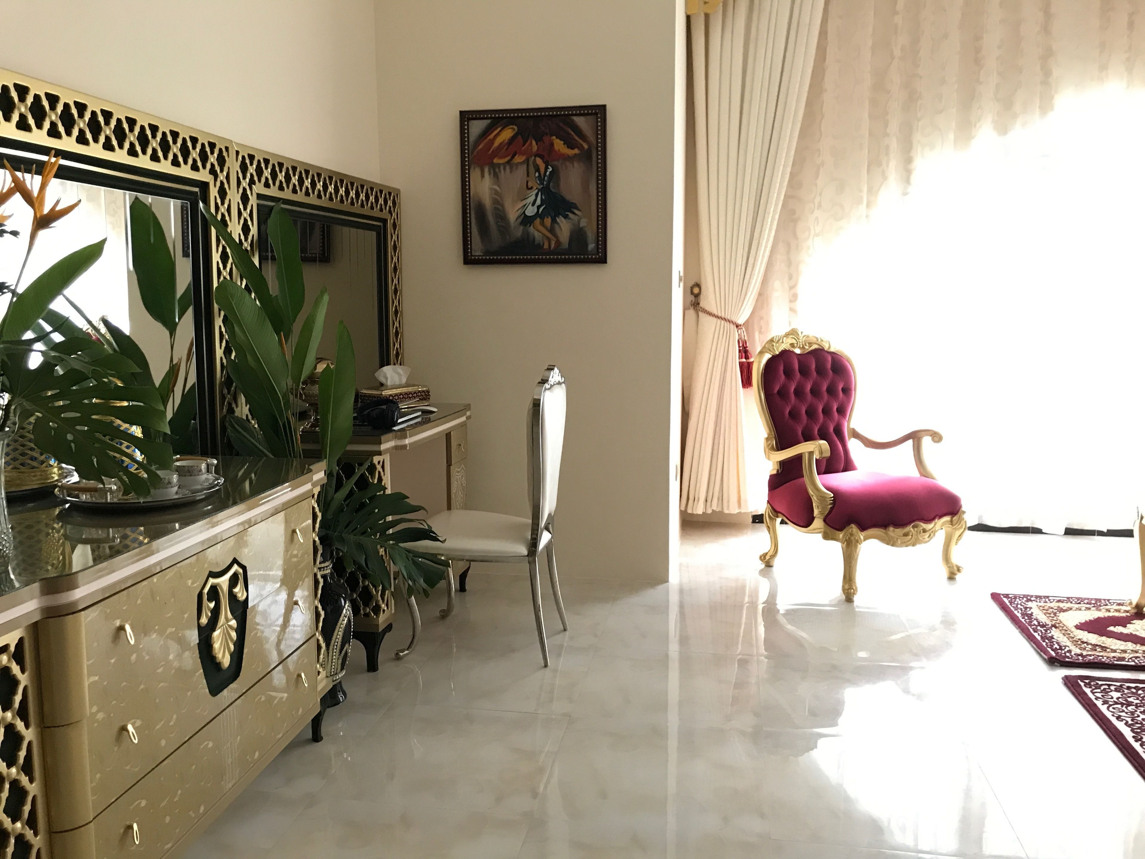 Een luxueuze kamer met een marmeren vloer, een kastje met gouden accenten, een zachte rode fauteuil en een grote spiegel met een decoratieve lijst.