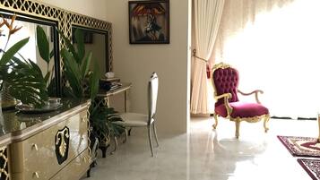 Een luxueuze kamer met een marmeren vloer, een kastje met gouden accenten, een zachte rode fauteuil en een grote spiegel met een decoratieve lijst.