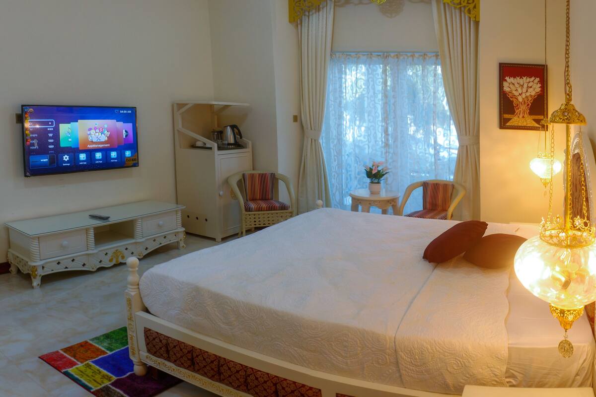 Une chambre d’hôtel avec un lit, un téléviseur fixé au mur, une petite table avec une chaise et un luminaire décoratif suspendu au plafond.