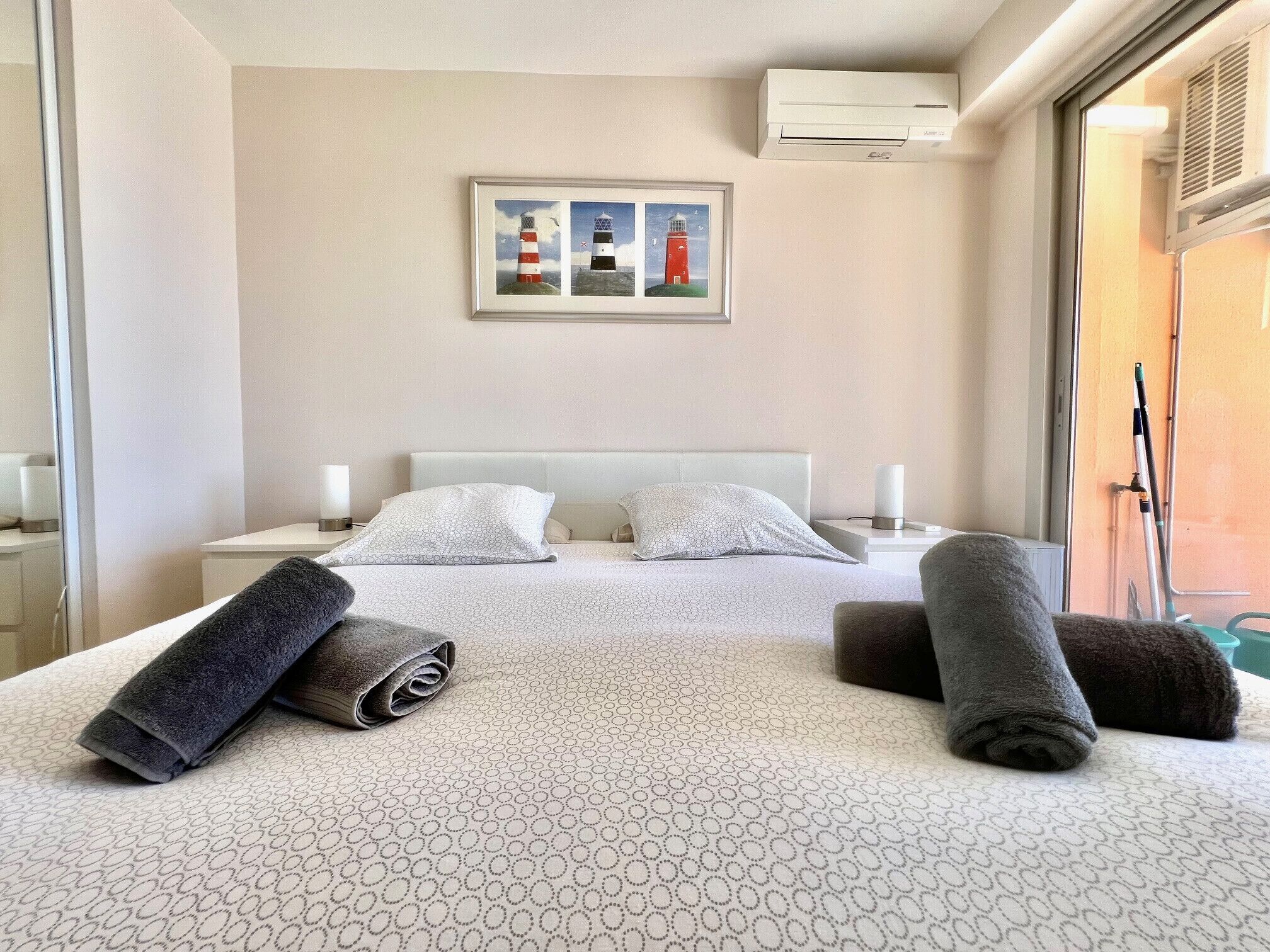 A hotel room with a bed, two pillows, a framed picture of lighthouses, a bedside table with a lamp, and a window with blinds.