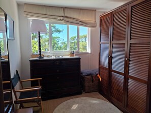 A room with a dresser, a lamp, a window with a view of trees, a chair, and a closet with louvered doors.