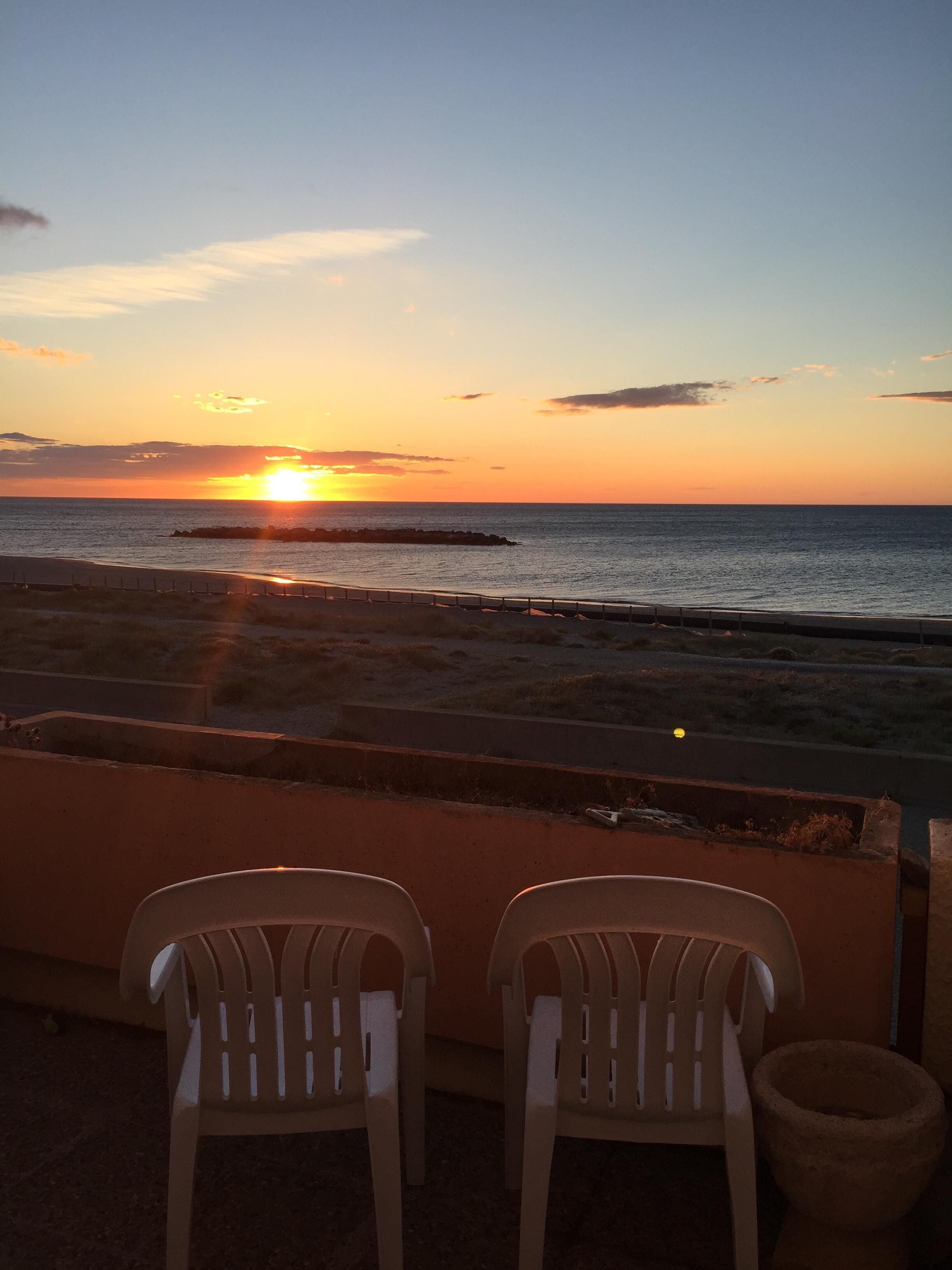 Une vue sur le coucher de soleil depuis un balcon, avec deux chaises en plastique et un bac à fleurs en pierre.