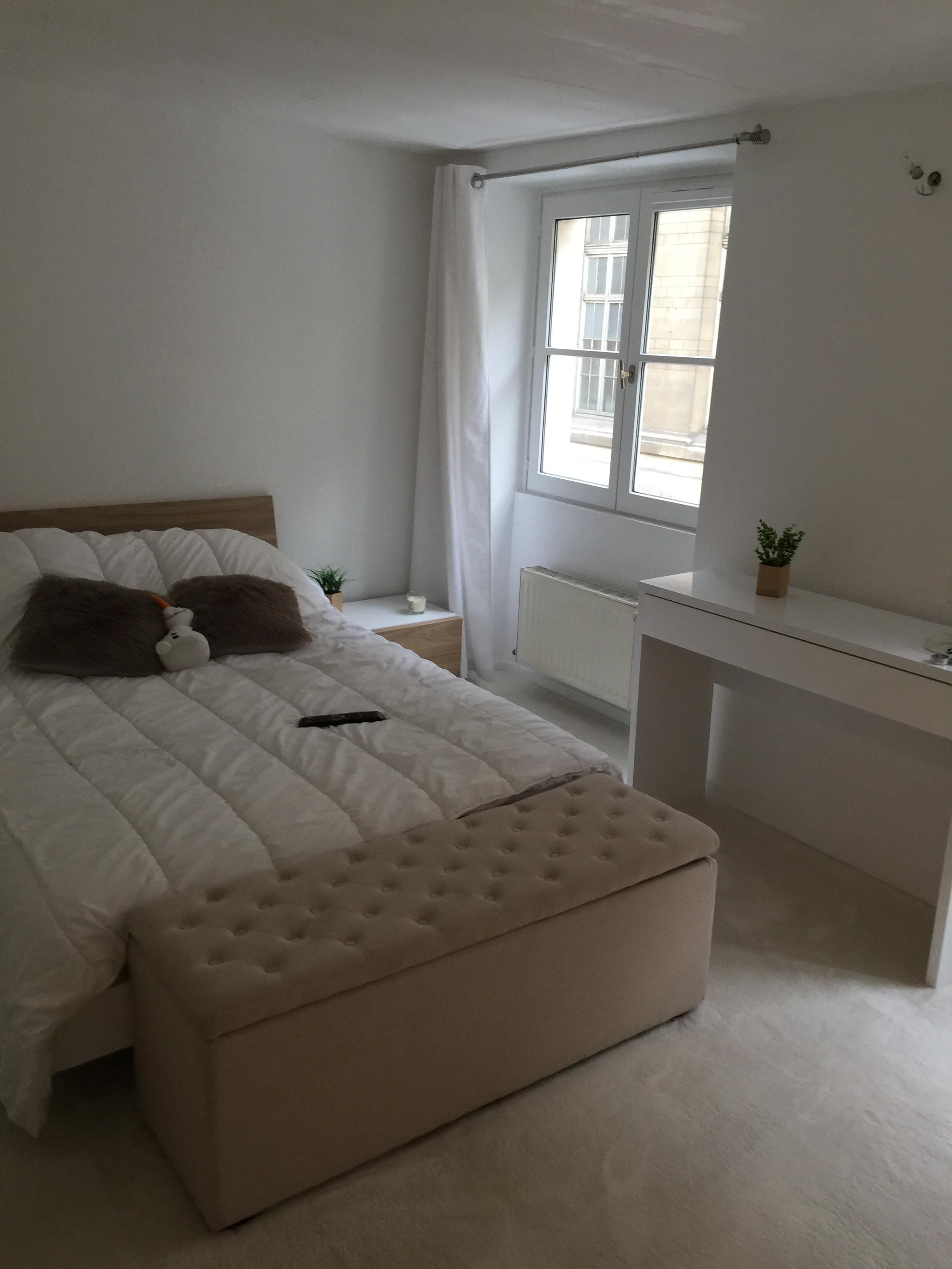 A bedroom with a bed, a bedside table, a radiator, a window with curtains, a small plant, and a desk.