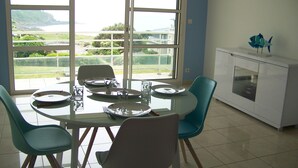 Ein Essbereich mit einem Tisch für vier Personen, Stühlen und Blick auf eine Küstenlandschaft durch große Fenster.