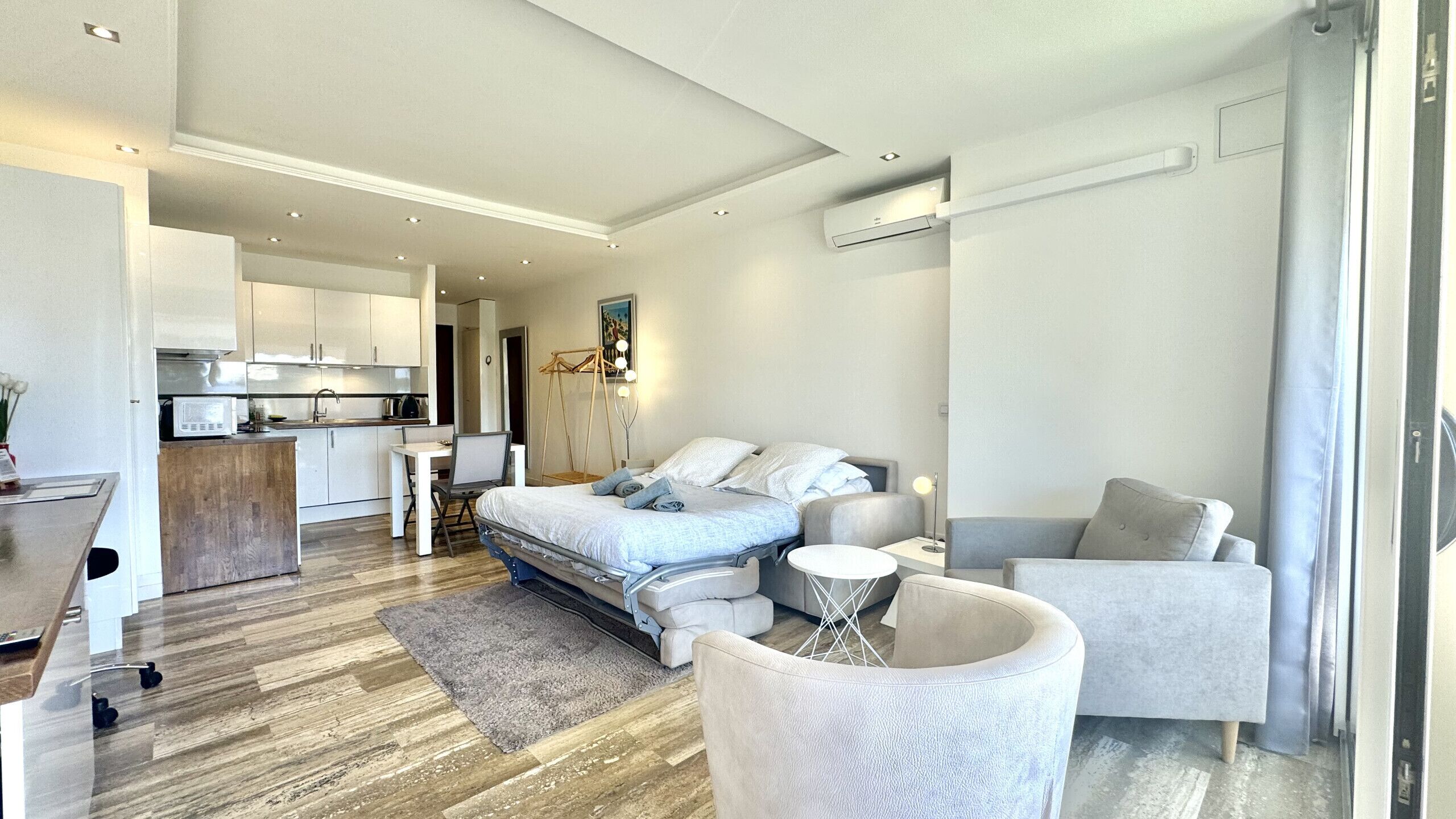 Un dormitorio moderno con una cama, dos sillones, una mesita y una kitchenette al fondo.