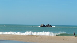 Una playa con un promontorio rocoso en el agua, olas rompiendo en la orilla y una persona de pie en la playa.