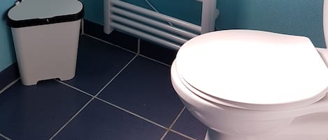 A bathroom with a white toilet, a trash bin, and a white towel rack against a teal wall.
