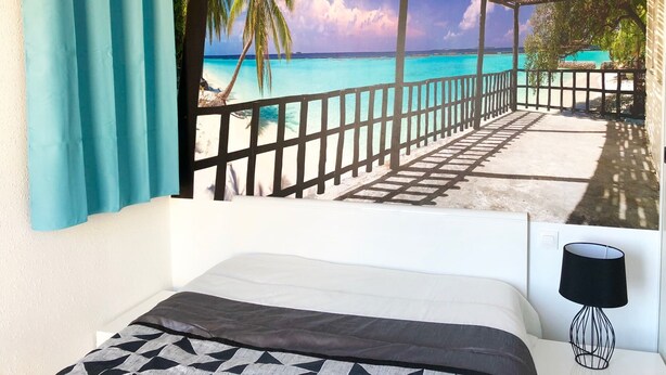 Een slaapkamer met een bed, een zwarte lamp en uitzicht op een strand door een raam met houten spijlen.