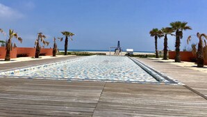 Een houten terras met een betegeld zwembad, palmbomen en een helder uitzicht op het strand.