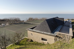 Uma quadra de tênis com vista para o mar, um edifício de pedra com telhado inclinado e uma área gramada.