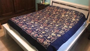 Ein Bett mit gemustertem Überwurf, ein Nachttisch mit Lampe sowie ein hölzerner Kleiderschrank in einem Zimmer mit hellblauen Wänden.