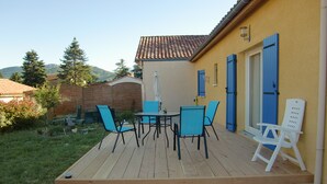 Un patio con piso de madera, mobiliario exterior y un edificio amarillo con contraventanas azules.