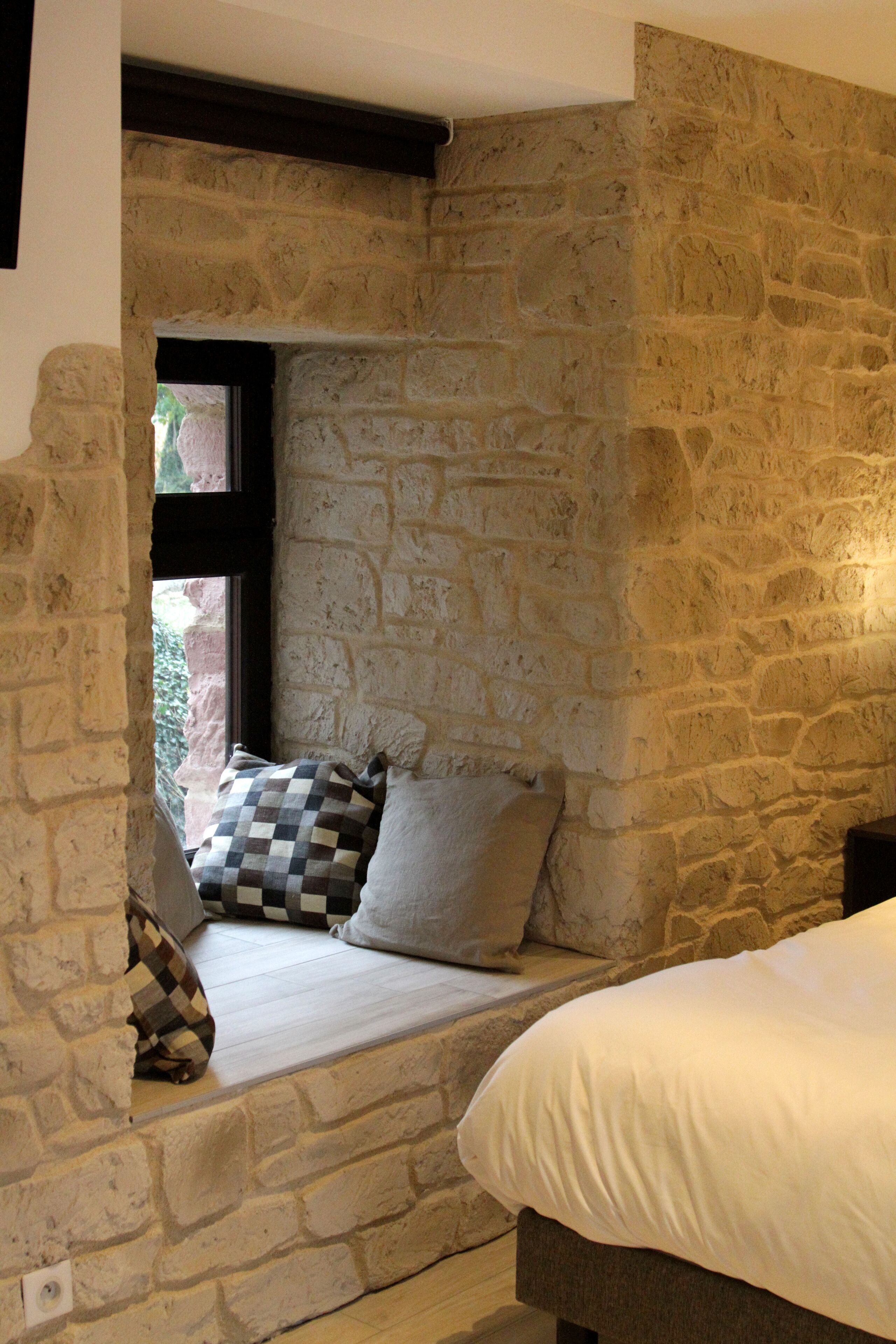 A stone-walled room with a window, a bed, and a checkered pillow.