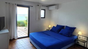 Ein Schlafzimmer mit einem Bett, zwei Nachttischen mit Lampen, einem Fernseher auf einem Schrank und Blick ins Freie durch eine Schiebetür.
