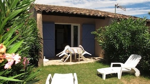 Uma casa com telhado de cerâmica, porta com persianas e um pequeno alpendre com duas poltronas brancas e uma mesa.