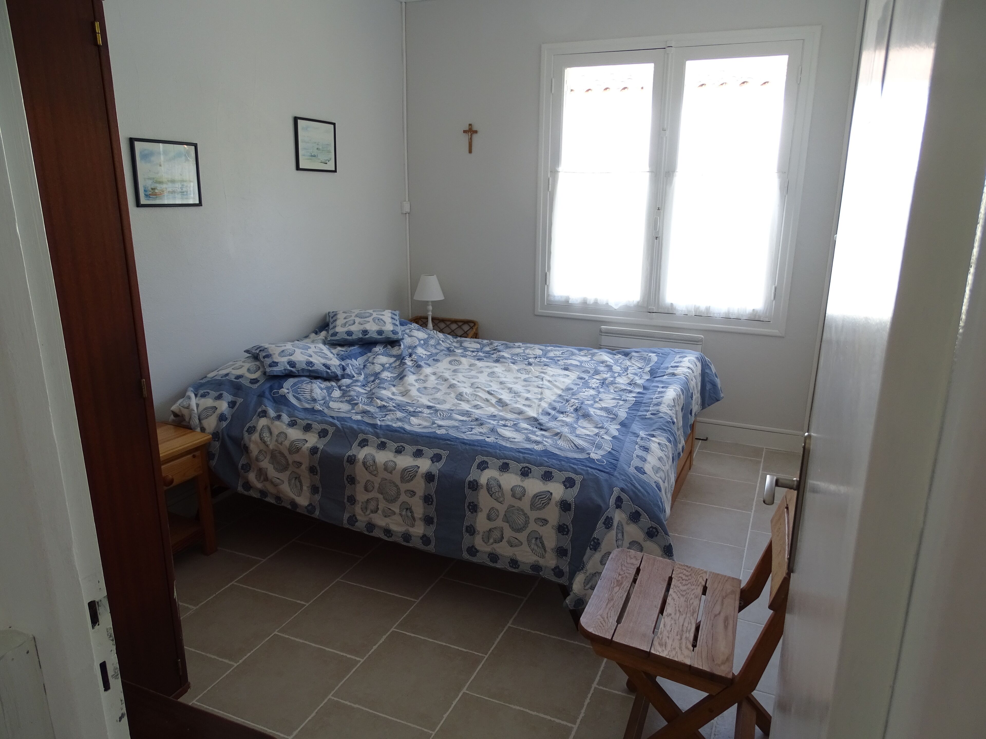 Un dormitorio con una cama, una silla de madera, una mesita de noche, una lámpara, una ventana y una cruz en la pared.