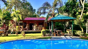 A pool with a wooden deck, a covered patio, and a building with a red roof.