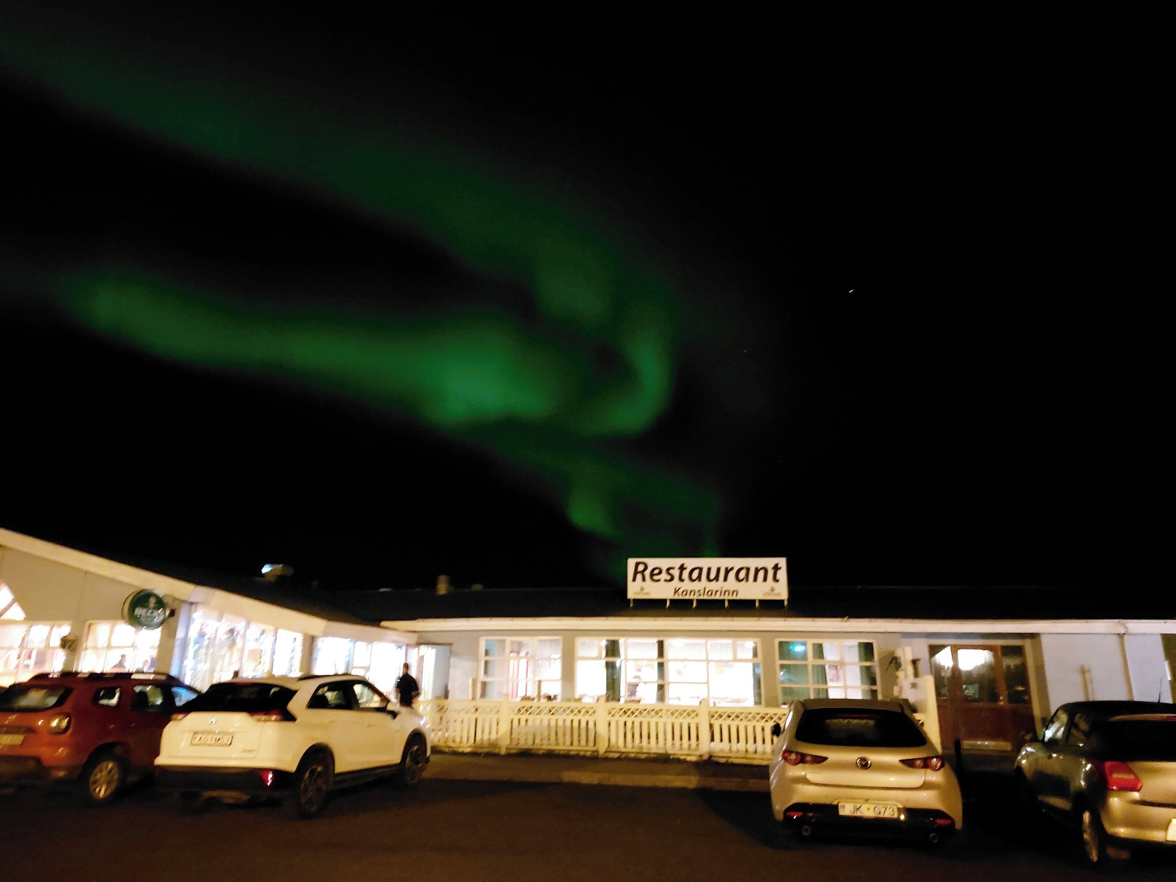 A restaurant with a sign, parked cars, and a view of the night sky with green lights.