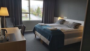 A hotel room with two beds, a desk with a lamp, and a view of the street through a window. - Hotel Kanslarinn (Rangárþing ytra)