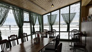 A dining area with wooden tables and chairs, large windows with green curtains, and a view of a snowy landscape. - Hotel Kanslarinn (Rangárþing ytra)