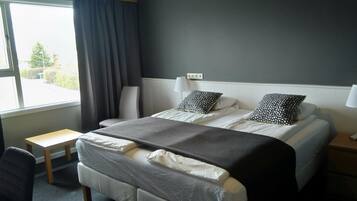 A hotel room with a bed, two pillows, a bedside table, a chair, and a window with curtains.