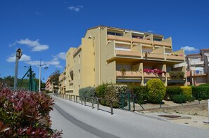 Un immeuble résidentiel à plusieurs étages, doté de balcons, avec un ciel dégagé et une rue bordée d’une clôture et de verdure.
