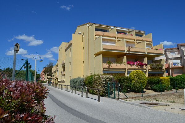 Un immeuble résidentiel à plusieurs étages, doté de balcons, avec un ciel dégagé et une rue bordée d’une clôture et de verdure.