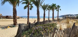 Une plage bordée de palmiers, une clôture en bois et une zone sablonneuse où sont entreposés des équipements de plage.