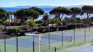 Una cancha de tenis con red, rodeada de árboles y vista al mar.