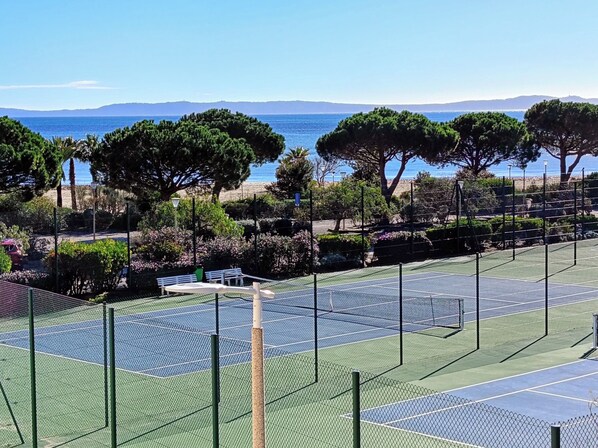 A tennis court with a net, surrounded by trees and a view of the sea.