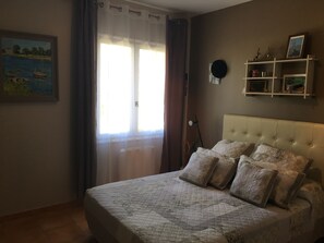 Een slaapkamer met een groot bed, een schilderij aan de muur, een raam met gordijnen en een plank met decoratieve objecten.