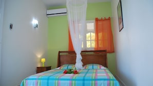 A bedroom with a bed, a bedside table with a lamp, a window with curtains, and an air conditioning unit on the wall. - Superb accommodation in a green location with private pool. (Sainte Anne)