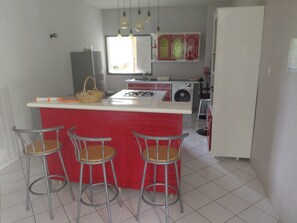 Une cuisine avec un îlot central rouge, quatre tabourets de bar, une cuisinière à gaz et des luminaires suspendus.