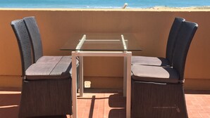 Een balkon met een glazen tafel en twee rieten stoelen, met uitzicht op een strand met de oceaan en een zeilboot in de verte.