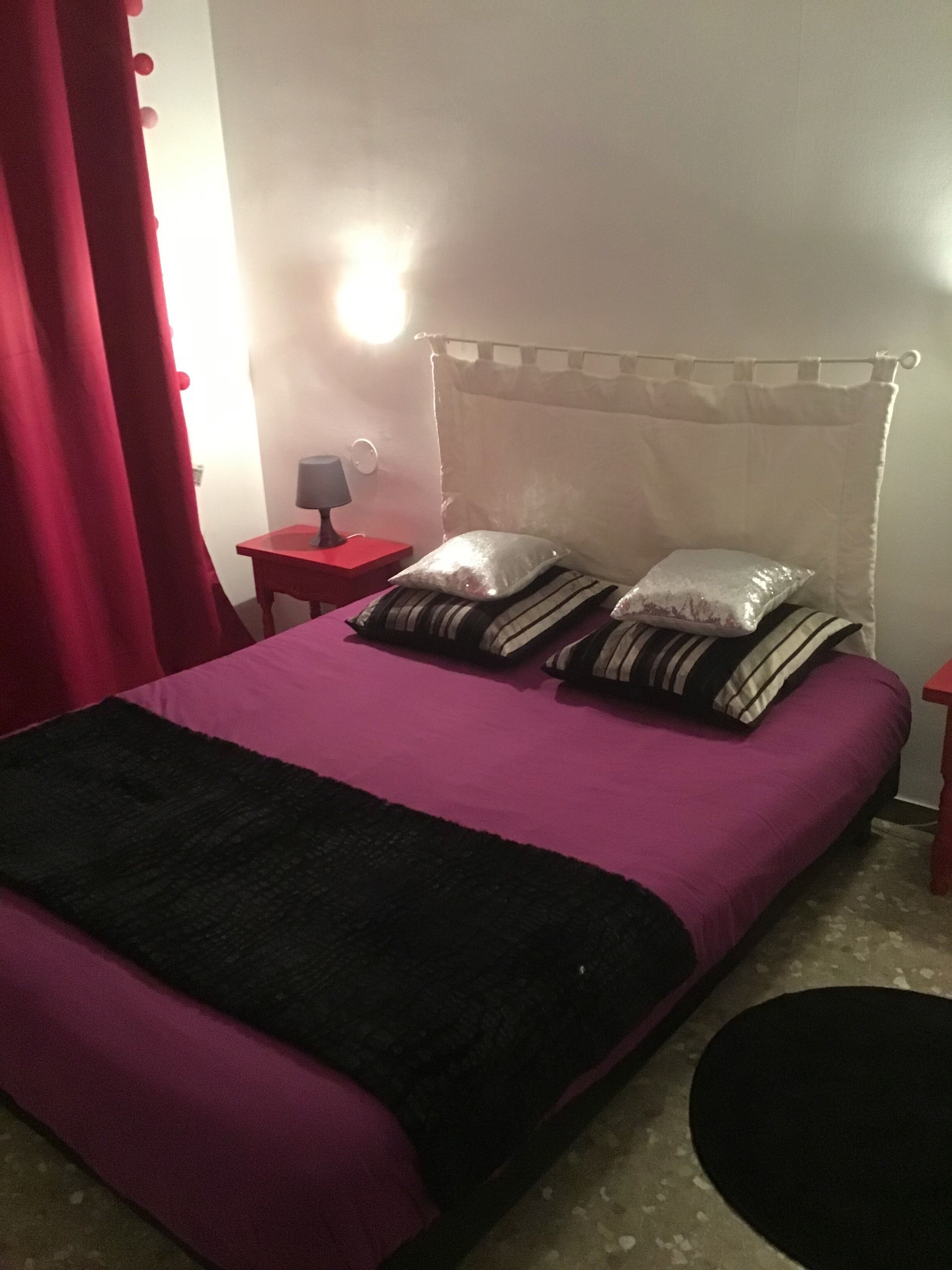 A bed with a purple bedspread and black and white striped pillows, a red nightstand with a lamp, and a white curtain.